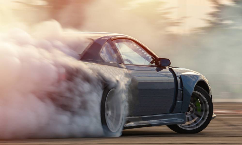 A dark-colored vehicle attempts to drift down an asphalt road. Smoke is billowing around the back tires.
