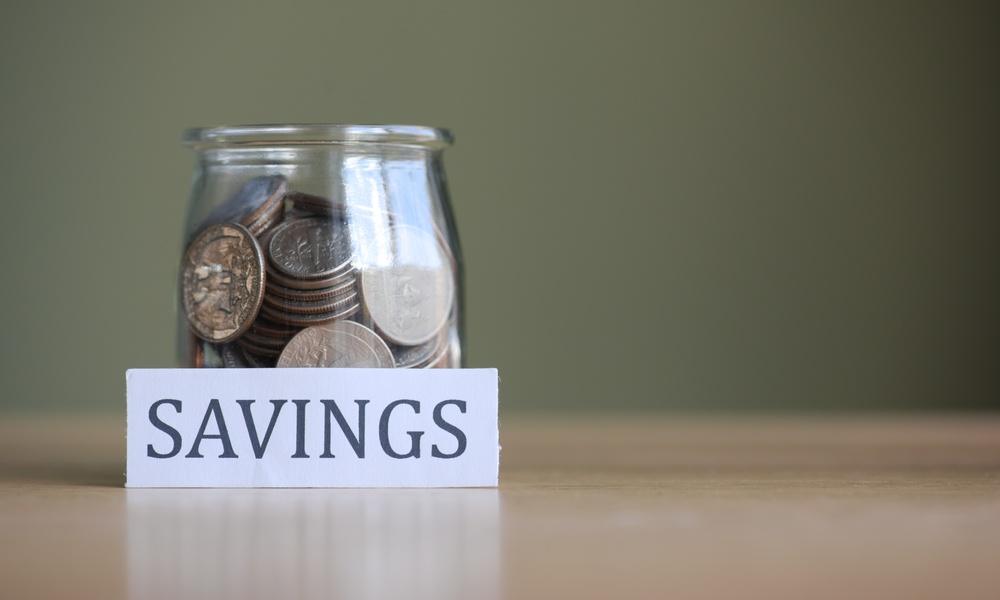 A jar of silver dollars on a plain table with a little note sitting in front of the jar labeled "savings."