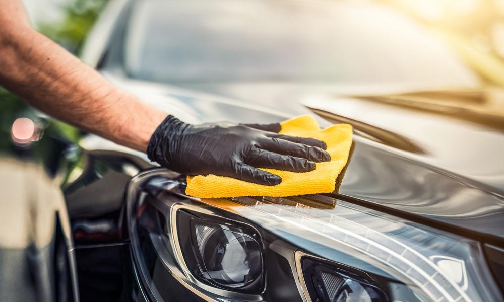 A close-up of a hand with latex gloves and a yellow cleaning rag wiping the headlight of a shining black car.