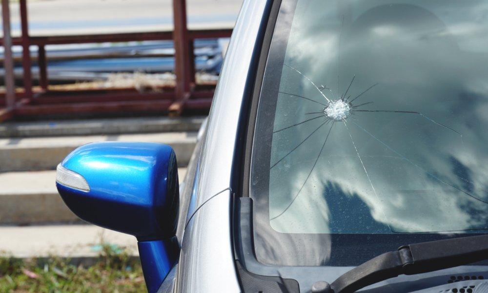 A silver car with a cracked windshield from a puncture in the bottom corner. The rear view mirror is blue.