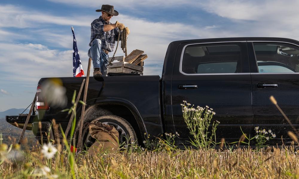 A man in a cowboy hat kneels in a black pickup bed, handling a rope beside a crate and an American flag.